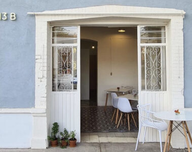 Entrada principal del primer local de Sunday Morning Café fachada de color azul con portón blanco y mesas de madera y melanina blanca